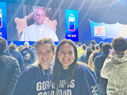 Rylee Miller, left, and Isabelle Bedel of St. Susanna Parish in Plainfield, Ind., pose for a photo as Pope Leo XIV appears on a huge screen in the background at Lucas Oil Stadium in Indianapolis on Nov. 21 during the National Catholic Youth Conference. (Photo by John Shaughnessy)